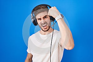 Hispanic man with beard listening to music wearing headphones angry and mad raising fist frustrated and furious while shouting