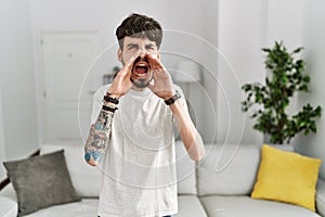 Hispanic man with beard at the living room at home shouting angry out loud with hands over mouth