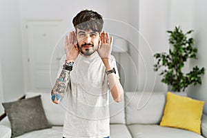 Hispanic man with beard at the living room at home trying to hear both hands on ear gesture, curious for gossip