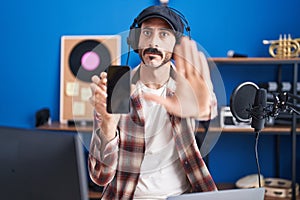 Hispanic man with beard at music studio showing smartphone with open hand doing stop sign with serious and confident expression,