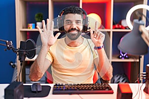 Hispanic man with beard playing video games with headphones showing and pointing up with fingers number seven while smiling