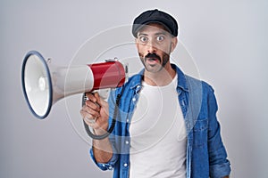 Hispanic man with beard shouting through megaphone scared and amazed with open mouth for surprise, disbelief face