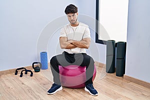 Hispanic man with beard sitting on pilate balls at yoga room skeptic and nervous, disapproving expression on face with crossed