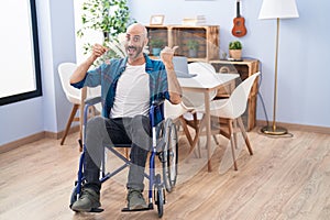 Hispanic man with beard sitting on wheelchair holding new house keys pointing thumb up to the side smiling happy with open mouth