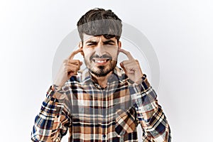 Hispanic man with beard standing over isolated background covering ears with fingers with annoyed expression for the noise of loud