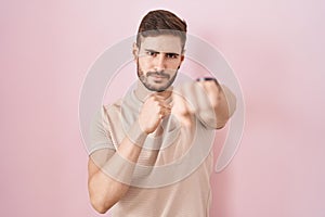 Hispanic man with beard standing over pink background punching fist to fight, aggressive and angry attack, threat and violence