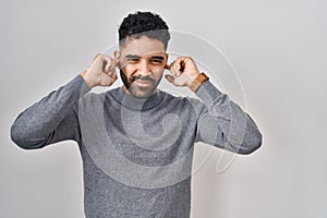 Hispanic man with beard standing over white background covering ears with fingers with annoyed expression for the noise of loud