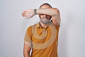Hispanic man with beard standing over white background covering eyes with arm, looking serious and sad