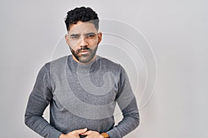 Hispanic man with beard standing over white background with hand on stomach because indigestion, painful illness feeling unwell