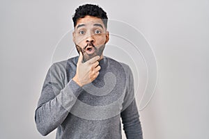 Hispanic man with beard standing over white background looking fascinated with disbelief, surprise and amazed expression with