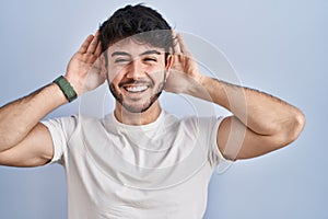 Hispanic man with beard standing over white background trying to hear both hands on ear gesture, curious for gossip