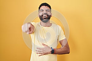 Hispanic man with beard standing over yellow background laughing at you, pointing finger to the camera with hand over body, shame