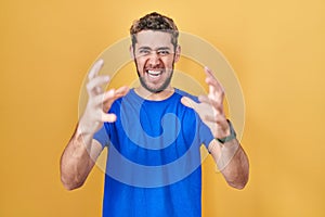 Hispanic man with beard standing over yellow background shouting frustrated with rage, hands trying to strangle, yelling mad