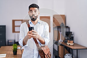 Hispanic man with beard using smartphone at the office making fish face with mouth and squinting eyes, crazy and comical
