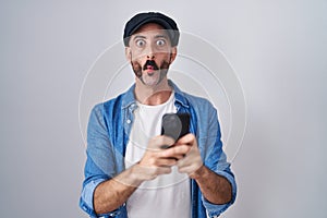 Hispanic man with beard using smartphone typing message making fish face with mouth and squinting eyes, crazy and comical