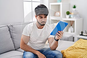 Hispanic man with beard using touchpad sitting on the sofa relaxed with serious expression on face