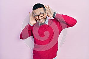 Hispanic man with beard wearing business shirt and glasses trying to hear both hands on ear gesture, curious for gossip