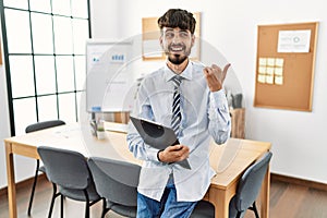 Hispanic man with beard wearing business style sitting on desk at office pointing thumb up to the side smiling happy with open