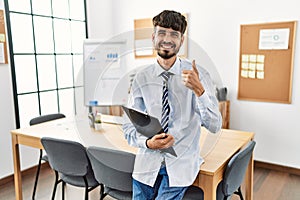 Hispanic man with beard wearing business style sitting on desk at office smiling happy and positive, thumb up doing excellent and