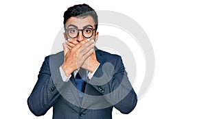 Hispanic man with beard wearing business suit and tie shocked covering mouth with hands for mistake