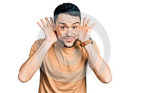 Hispanic man with beard wearing casual t shirt trying to hear both hands on ear gesture, curious for gossip
