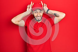 Hispanic man with beard wearing delivery uniform and cap doing bunny ears gesture with hands palms looking cynical and skeptical
