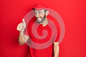 Hispanic man with beard wearing delivery uniform and cap looking proud, smiling doing thumbs up gesture to the side