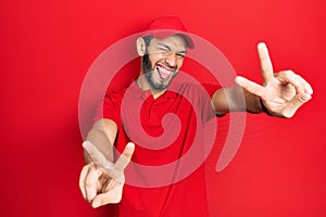 Hispanic man with beard wearing delivery uniform and cap smiling with tongue out showing fingers of both hands doing victory sign