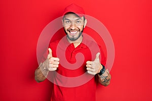 Hispanic man with beard wearing delivery uniform and cap success sign doing positive gesture with hand, thumbs up smiling and