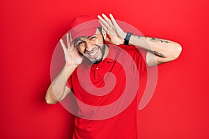Hispanic man with beard wearing delivery uniform and cap trying to hear both hands on ear gesture, curious for gossip