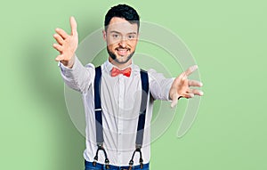 Hispanic man with beard wearing hipster look with bow tie and suspenders looking at the camera smiling with open arms for hug