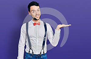 Hispanic man with beard wearing hipster look with bow tie and suspenders smiling cheerful presenting and pointing with palm of