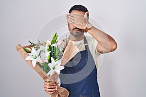 Hispanic man with beard working as florist covering eyes with hand, looking serious and sad