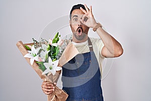 Hispanic man with beard working as florist doing ok gesture shocked with surprised face, eye looking through fingers