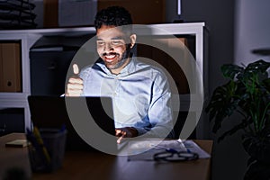 Hispanic man with beard working at the office with laptop at night smiling happy and positive, thumb up doing excellent and