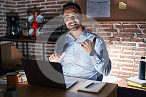 Hispanic man with beard working at the office at night pointing to the back behind with hand and thumbs up, smiling confident