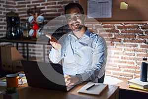 Hispanic man with beard working at the office at night smiling cheerful offering palm hand giving assistance and acceptance