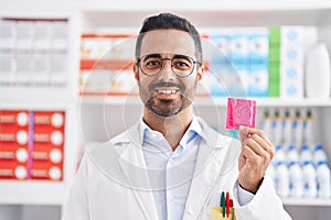 Hispanic man with beard working at pharmacy drugstore holding condom looking positive and happy standing and smiling with a