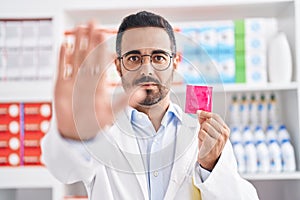 Hispanic man with beard working at pharmacy drugstore holding condom with open hand doing stop sign with serious and confident