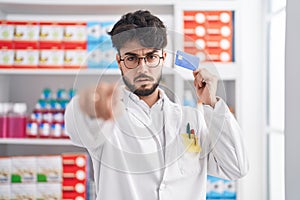 Hispanic man with beard working at pharmacy drugstore holding credit card pointing with finger to the camera and to you, confident