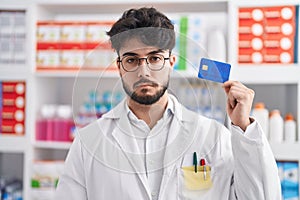 Hispanic man with beard working at pharmacy drugstore holding credit card thinking attitude and sober expression looking self