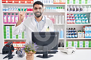 Hispanic man with beard working at pharmacy drugstore showing and pointing up with fingers number four while smiling confident and