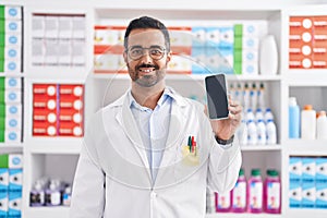 Hispanic man with beard working at pharmacy drugstore showing smartphone screen looking positive and happy standing and smiling