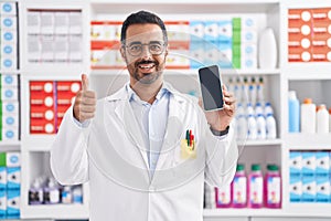 Hispanic man with beard working at pharmacy drugstore showing smartphone screen smiling happy and positive, thumb up doing