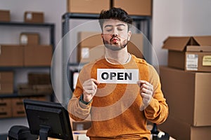 Hispanic man with beard working at small business ecommerce holding open sign looking at the camera blowing a kiss being lovely