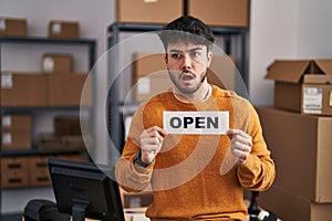 Hispanic man with beard working at small business ecommerce holding open sign in shock face, looking skeptical and sarcastic,