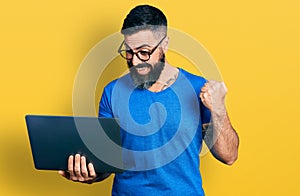 Hispanic man with beard working using computer laptop screaming proud, celebrating victory and success very excited with raised