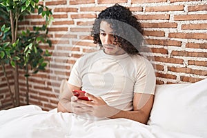 Hispanic man with curly hair using smartphone sitting on the bed scared and amazed with open mouth for surprise, disbelief face