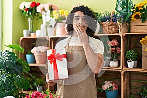 Hispanic man with curly hair working at florist shop holding gift covering mouth with hand, shocked and afraid for mistake