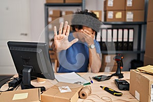 Hispanic man with curly hair working at small business ecommerce covering eyes with hands and doing stop gesture with sad and fear